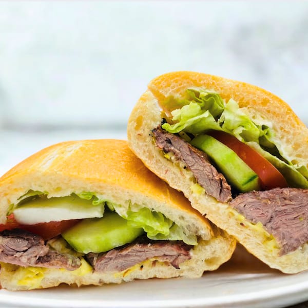 Two halves of a steak sandwich with lettuce, tomato, and avocado on a white plate.