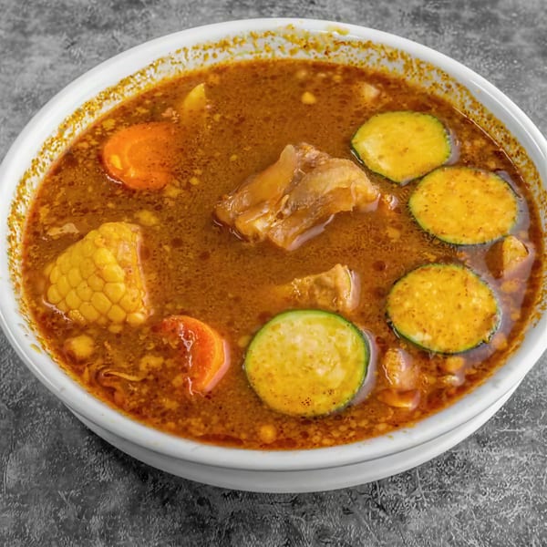 Hearty beef soup with corn on the cob, carrots, and zucchini slices in a white bowl.