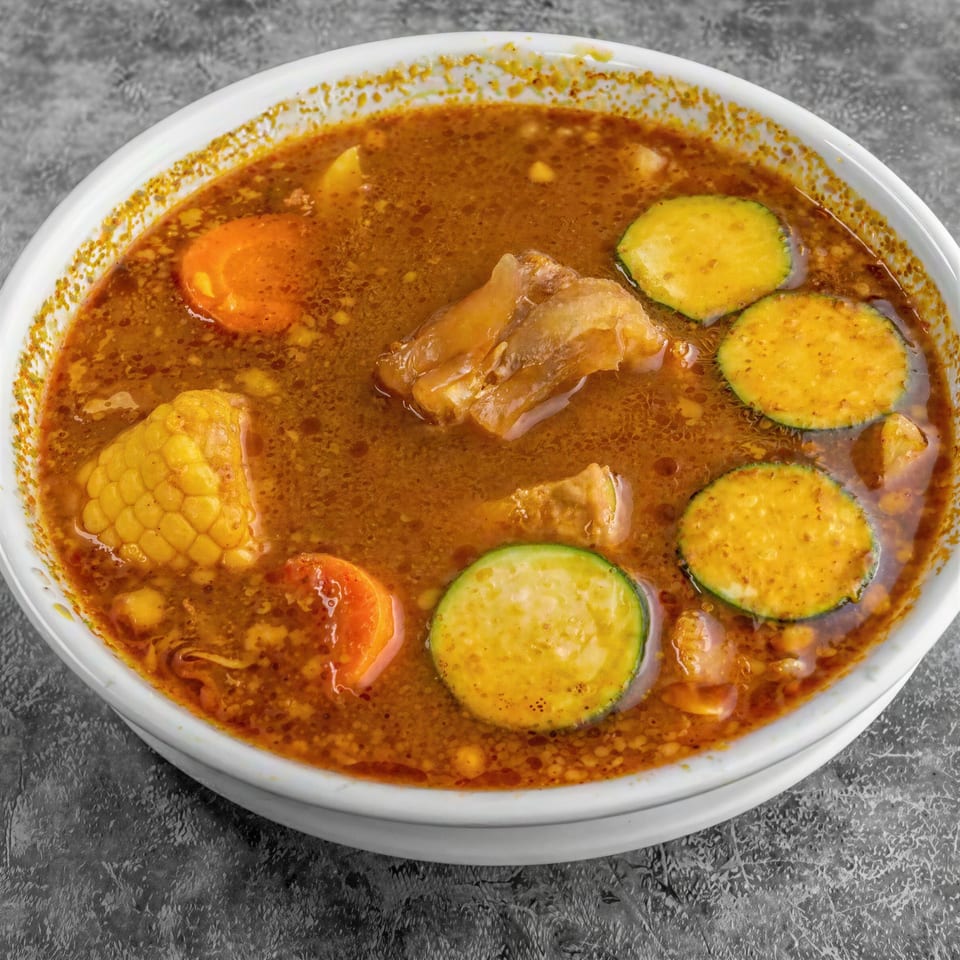 Hearty beef soup with corn on the cob, carrots, and zucchini slices in a white bowl.
